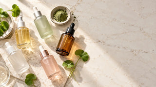 Hero editorial Korean serum flatlay featuring assorted frosted glass bottles, centella leaf, and green tea powder on white marble
